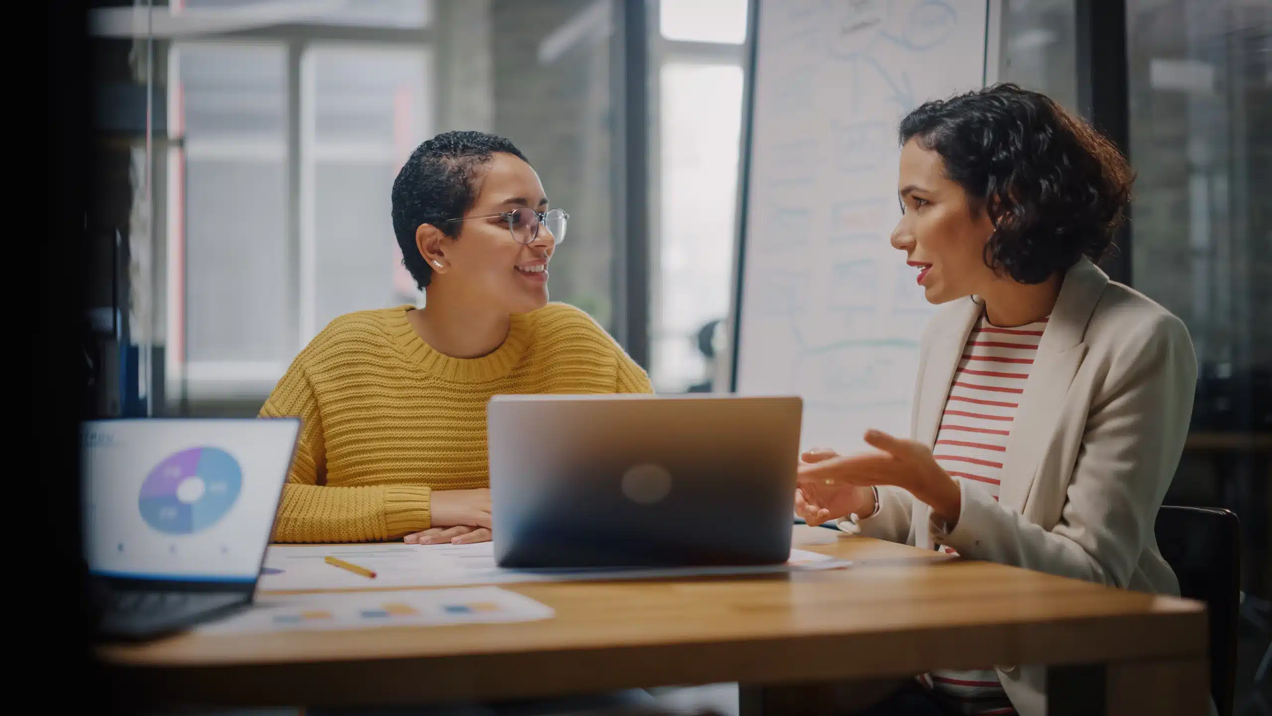 Two Diverse Multiethnic Female Have a Discussion in Meeting Room Behind Glass Walls in an Agency. Creative Director and Project Manager Compare Business Results on Laptop and App Designs in an Office. Two Latina women in a business meeting, representing authentic connection with Hispanic audiences in marketing